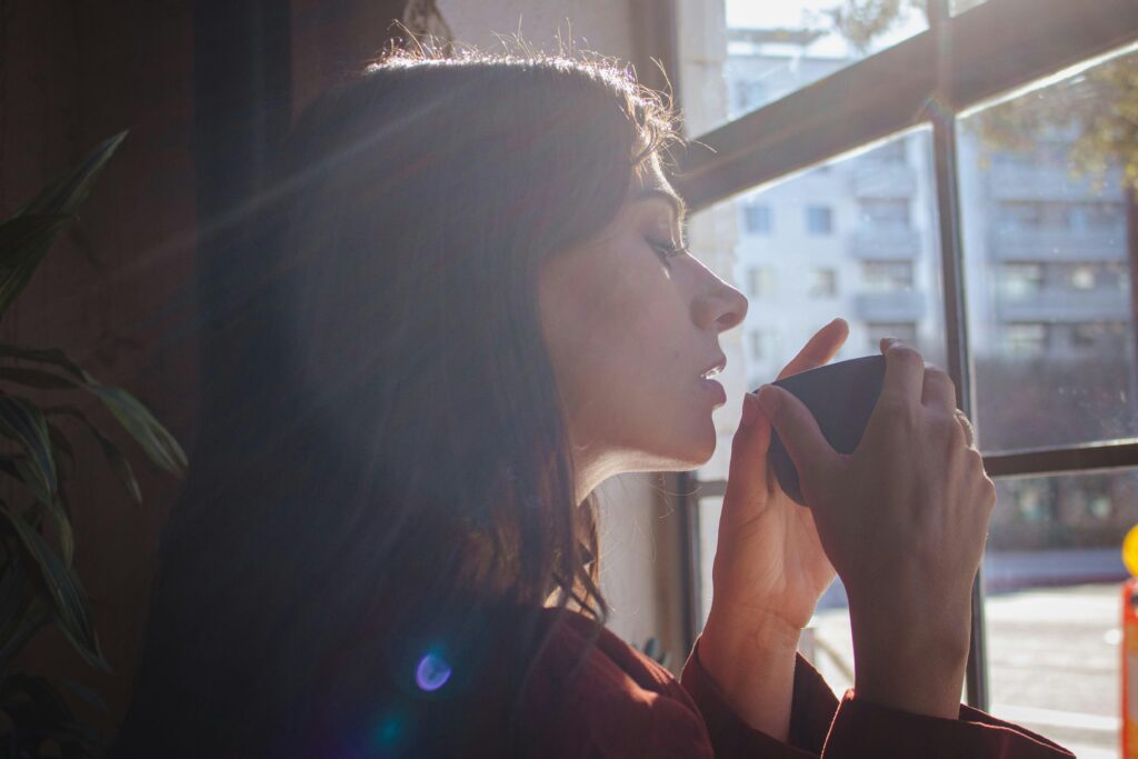 A woman savors a hot drink by a sunlit window in an urban setting.