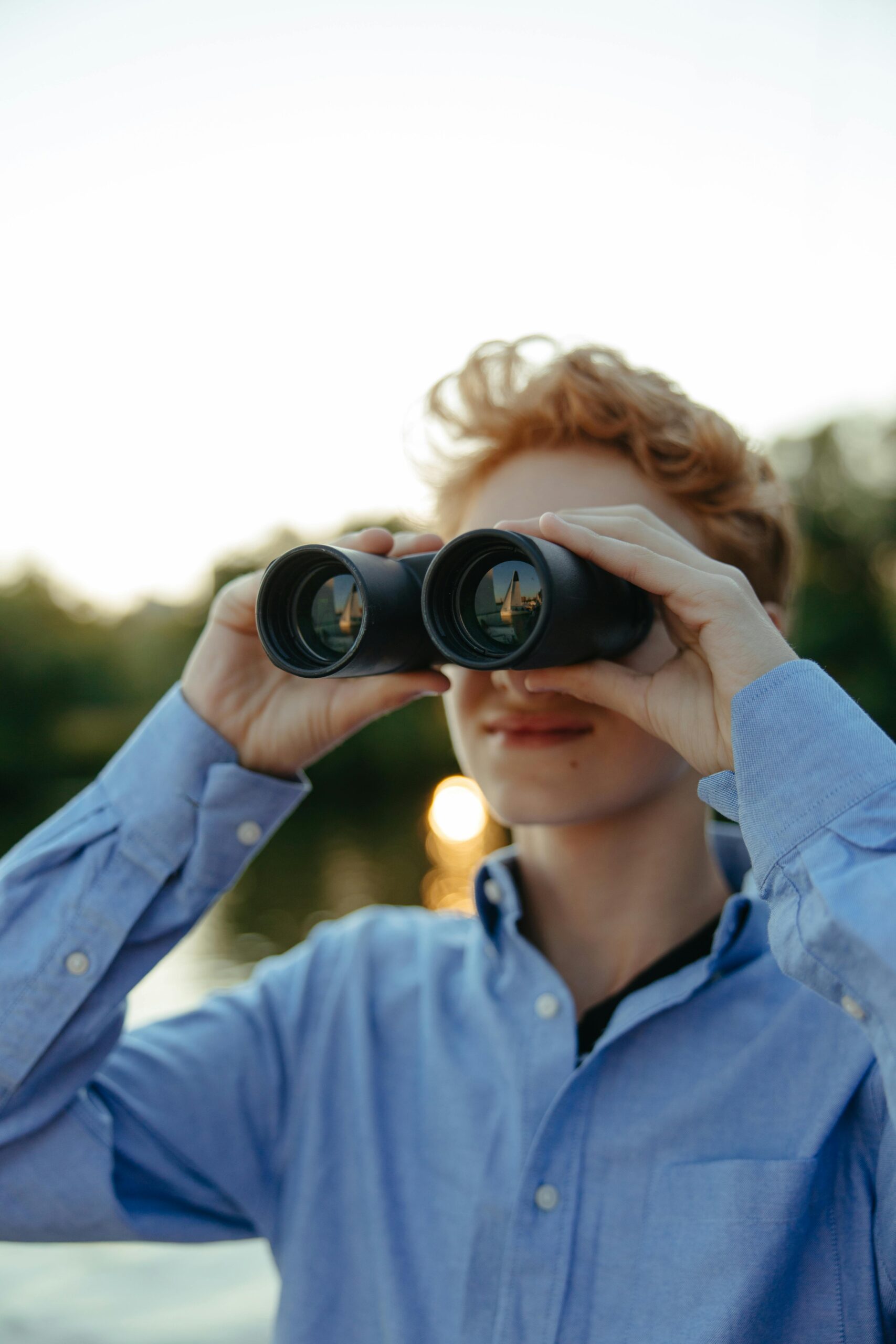 A young teenager in a blue shirt observes the scenery using binoculars during sunset.